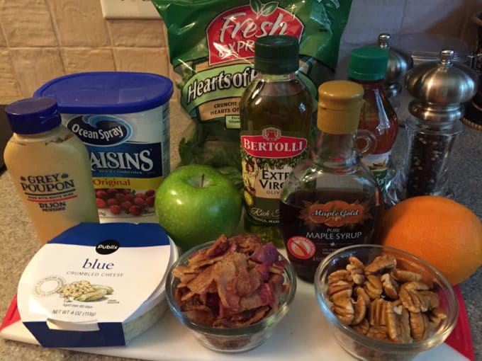 A container of blue cheese, cooked bacon, pecan halves, olive oil and maple syrup on a cutting board.