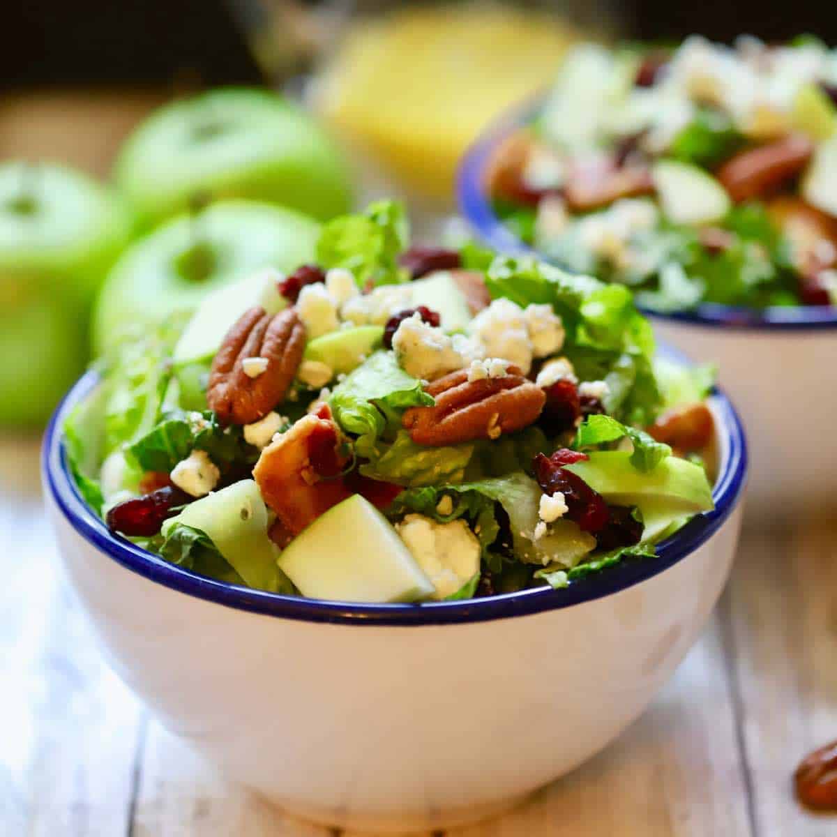 A large white bowl full of chopped salad topped with apples and pecans.