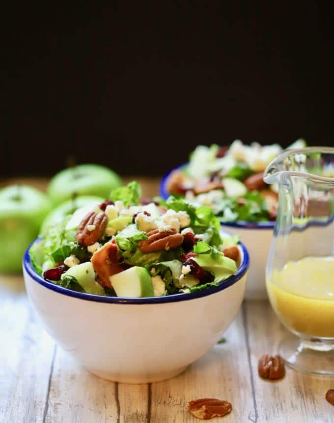 Two bowls of apple salad with green apples in the background and a small pitcher of salad dressing.