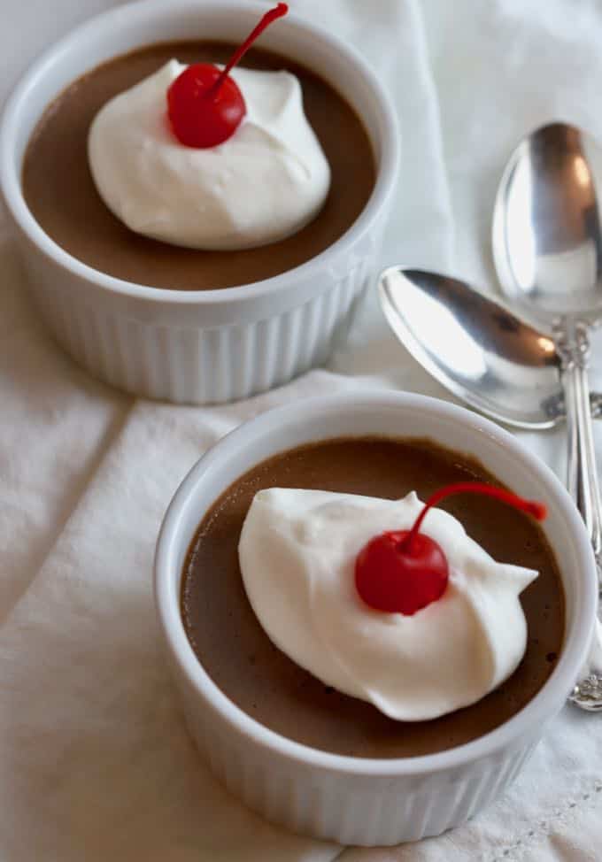 Two white bowls of chocolate pudding topped with whipped cream and a cherry.