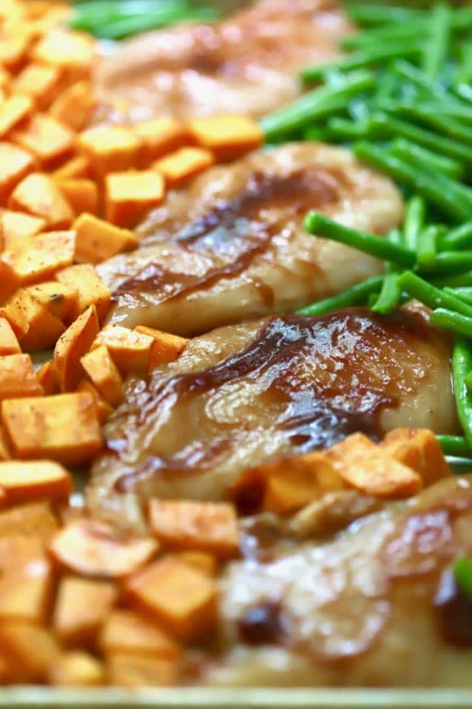 A sheet pan with sweet potato cubes, chicken breasts covered with BBQ sauce and green beans.