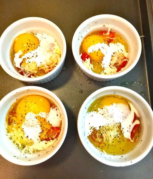 The four ramekins topped with an egg, seasoning, and heavy cream on a baking sheet.