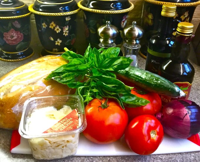 Tomatoes, cucumbers, french bread, parmesan cheese, and basil on a cutting board to make a salad. 