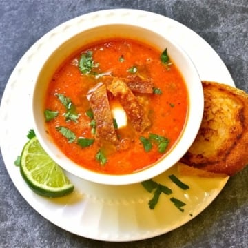 Tomato tortilla soup in a white bowl garnished with lime, tortilla chips and cilantro.