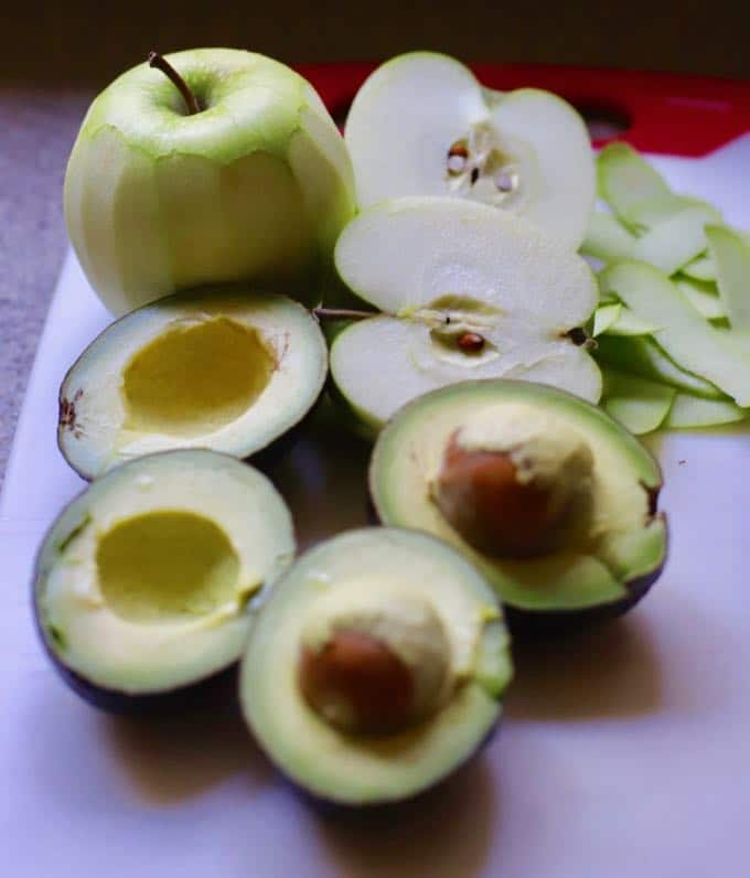 on a cutting board is an apple cut in half, one skinned apple, and two avocados cut in half.