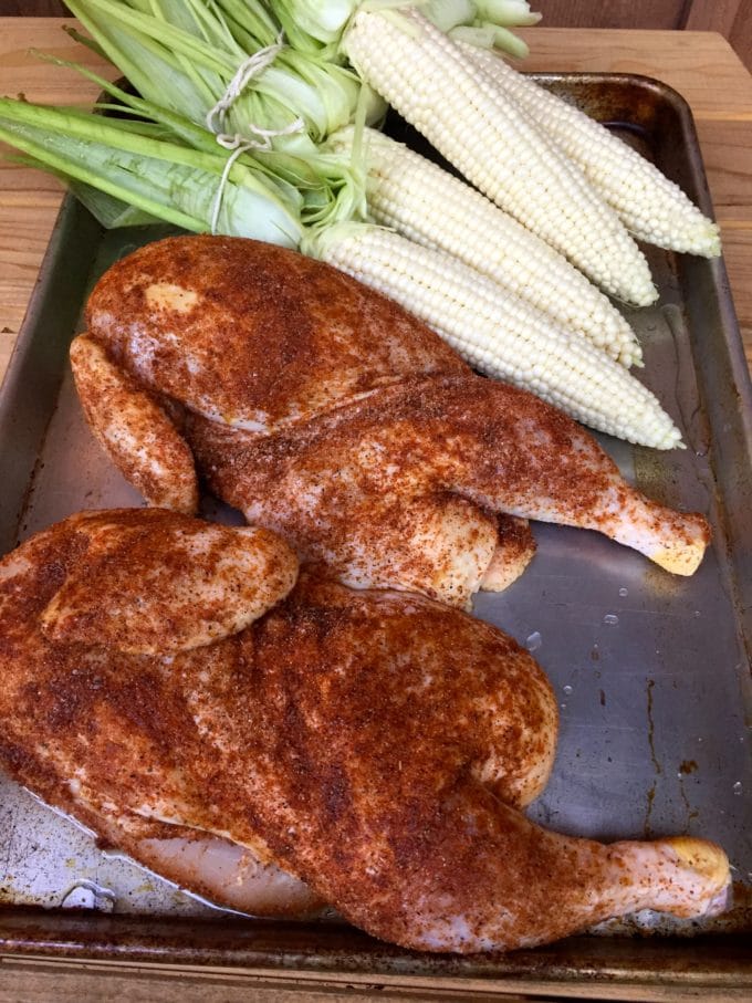Two chicken halves covered with a rub and corn ready to go on the grill. 
