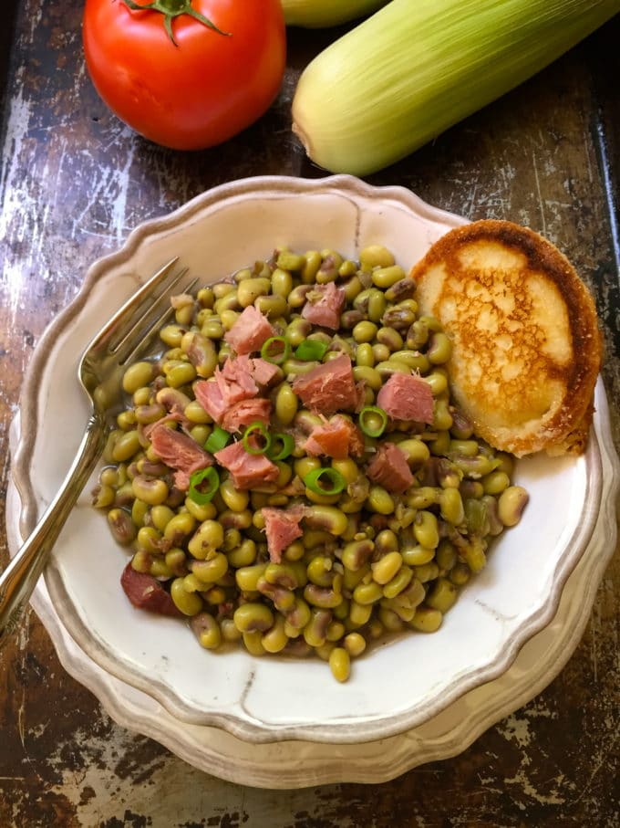 Southern peas in a white bowl topped with chunks of ham and a hoecake.