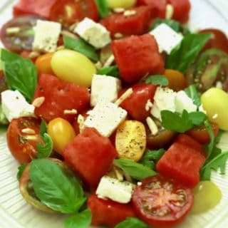 A large white plate filled with southern watermelon tomato salad garnished with feta cheese, slivered almonds, and chopped basil.