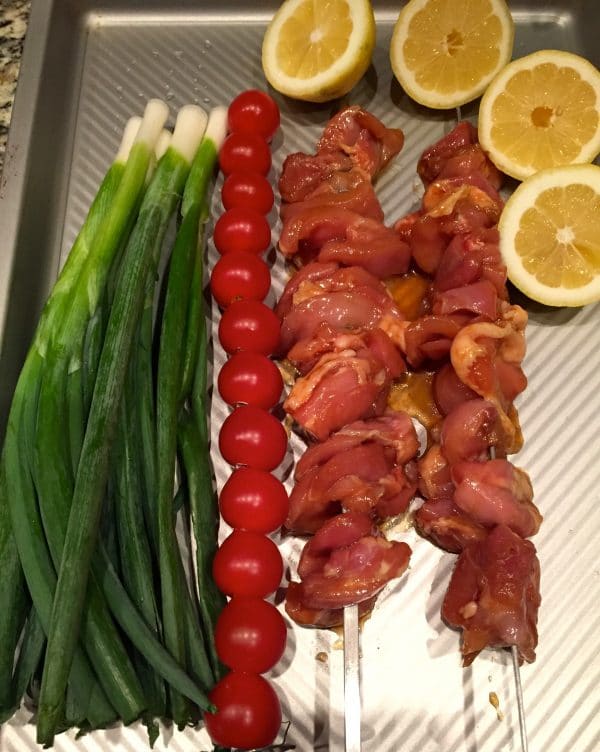 Chicken skewers, cherry tomato skewer, lemon halves, and scallions on a baking sheet.