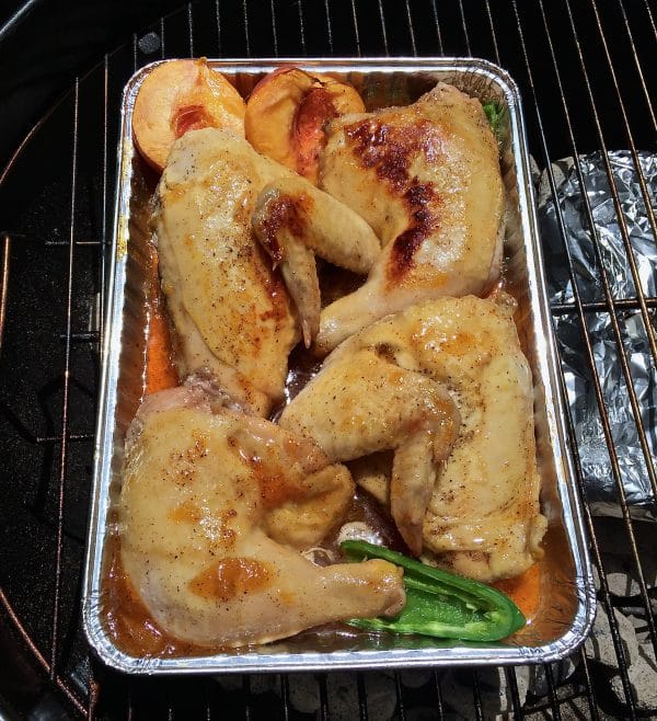 Chicken pieces in the pan with peach halves and jalapeño halves on the grill.