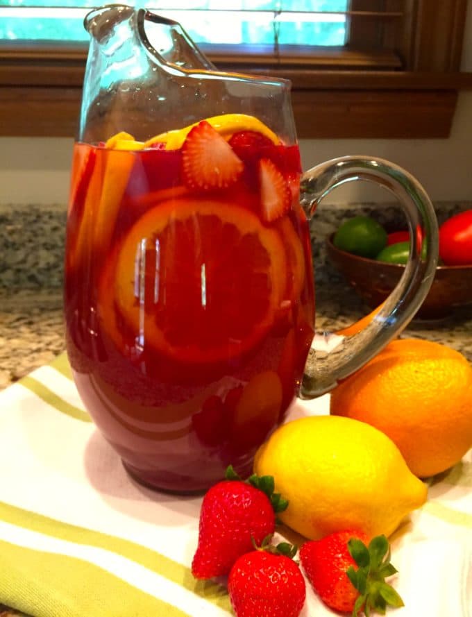 A tall glass pitcher of sangria garnished with strawberries, lemons and oranges