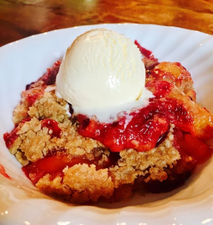 A Peach and Blackberry Crumble serving topped with vanilla ice cream in a white bowl.