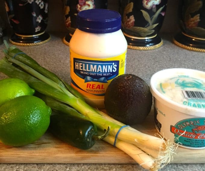 Two limes, a jalapeno pepper, scallions, a container of blue crab and a jar of mayonnaise on a cutting board..