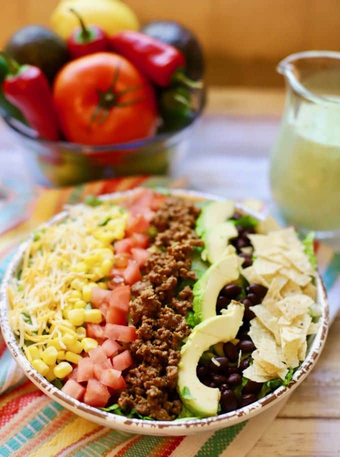 A bowl of vegetables next to a large taco salad and a pitcher of avocado dressing.