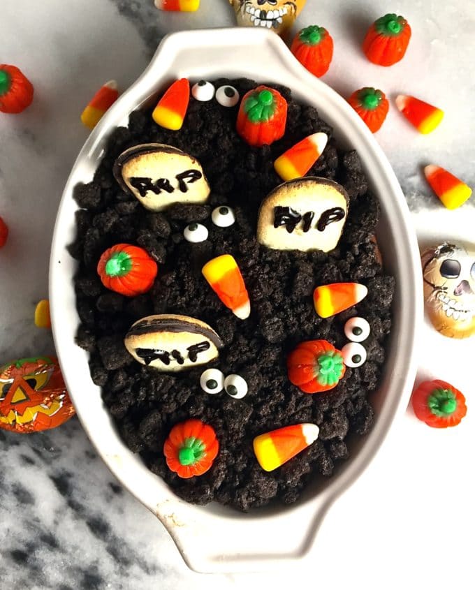 Graveyard Chocolate Cheesecake Dip in a white dish topped with a layer of crushed Oreos and tombstone cookies with RIP piped on with icing. 