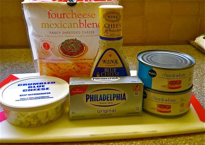 Buffalo Chicken Dip ingredients on a cutting board including crumbled blue cheese and canned chunk chicken.