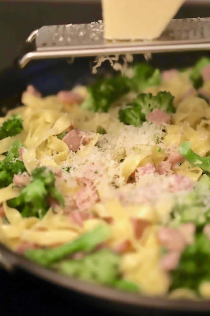 Grating parmesan cheese over Fettuccine Alfredo with Broccoli and Canadian Bacon in a large skillet.
