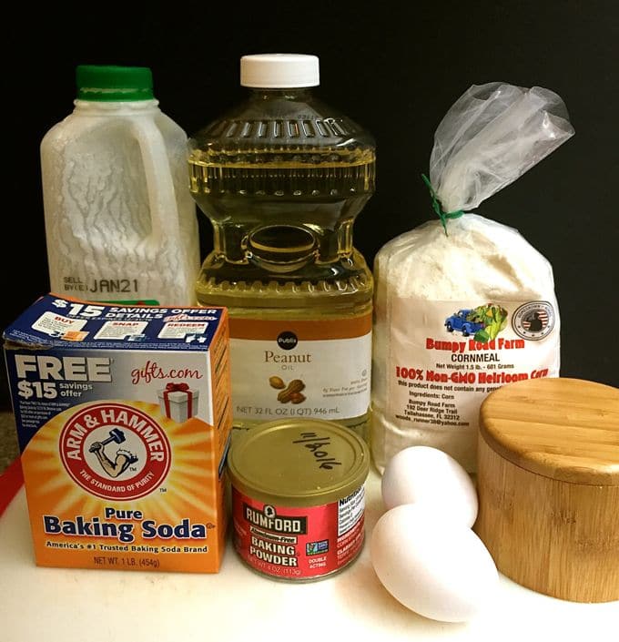 Baking soda, baking powder, two eggs, cornmeal, buttermilk and peanut oil on a table. 