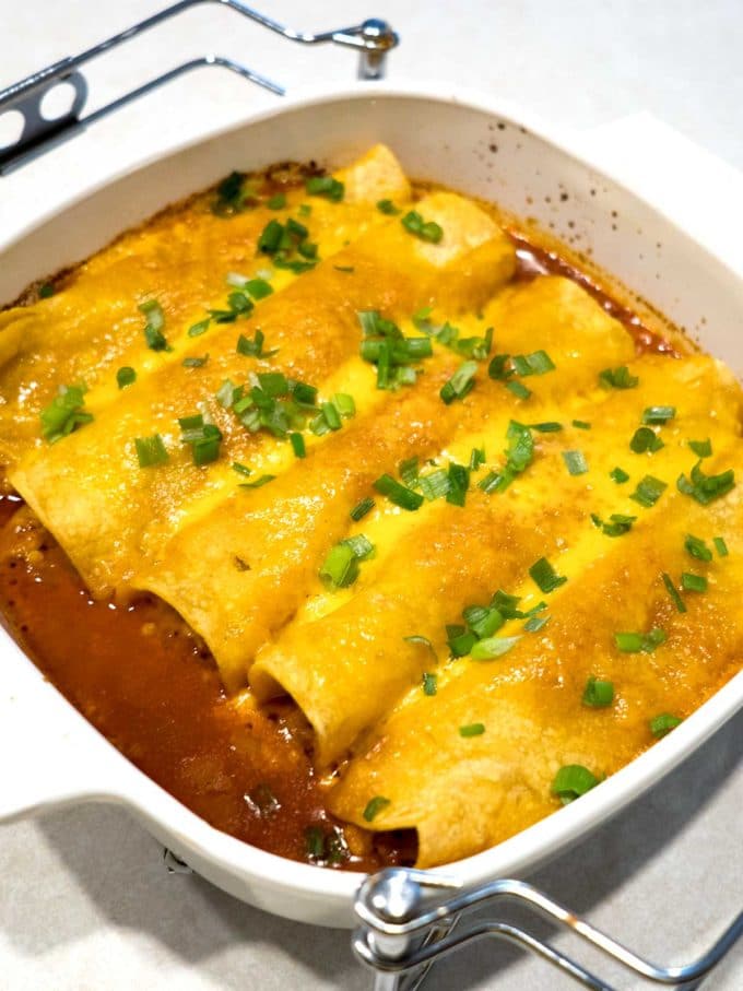 Chicken Enchiladas with Red Chili Sauce in a white baking dish topped with melted cheese and chopped chives