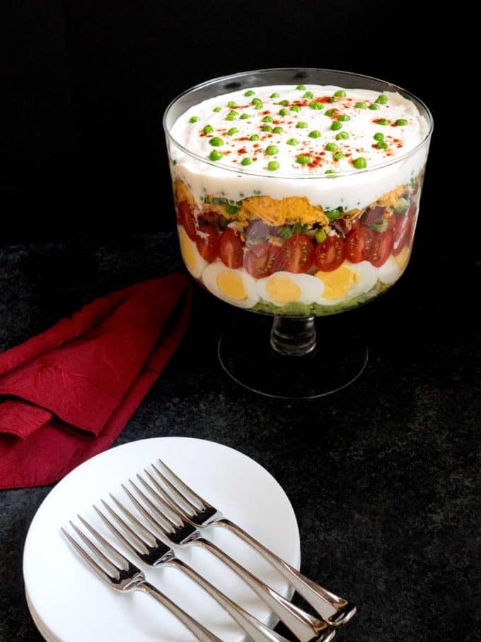A Seven Layer Salad in a clear glass trifle dish against a black background, with red napkin and white salad plates and forks.