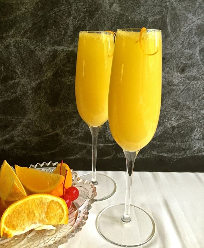 Two Classic Mimosas with a Twist on a counter with fresh orange slices in a bowl.