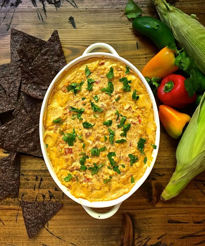 Easy Cheesy Hot Corn Dip on a wooden board surrounded by chips, peppers and ears of corn
