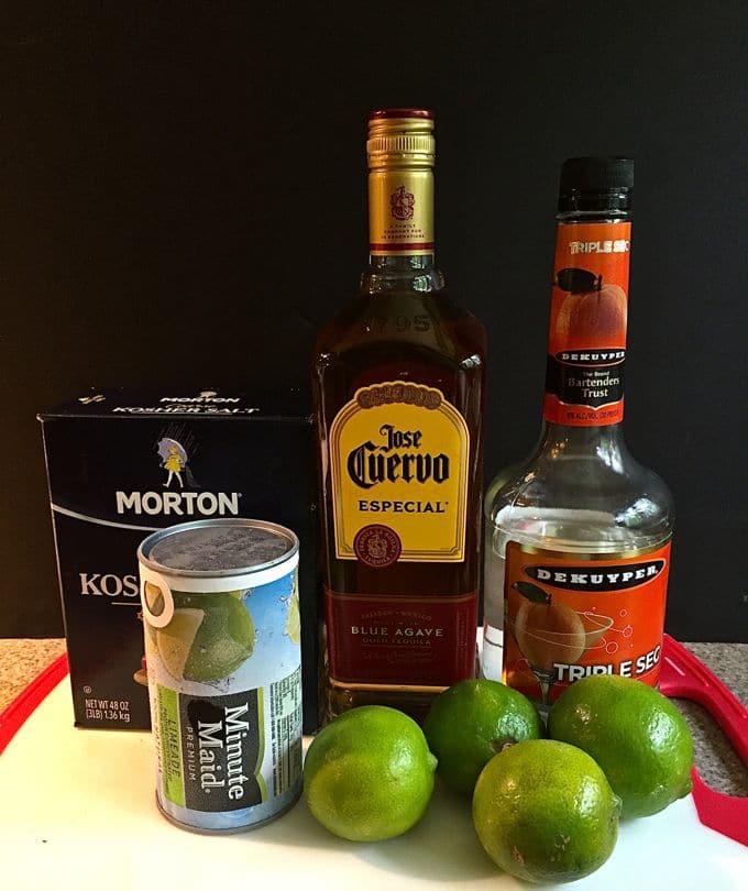 Margarita ingredients including tequila, triple sec, limeade and limes on a kitchen counter. 