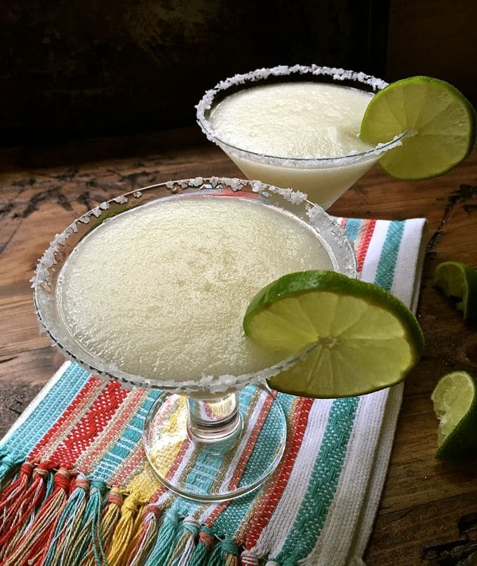 Two glasses full of frozen margaritas garnished with lime slices on a pretty Mexican towel. 