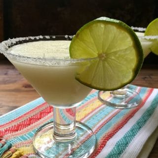 Frozen margaritas in margarita glasses on a colorful Mexican towel, garnished with lime slices.