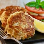 Three crab cakes on a black plate with a fork, sliced tomatoes and a slice of lemon.