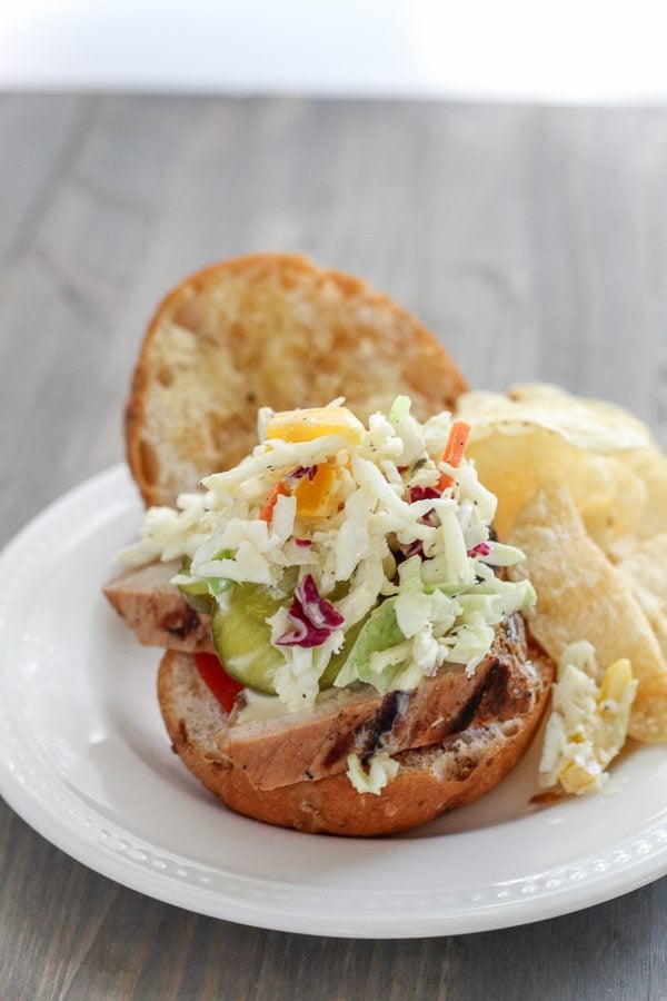 A grilled pork tenderloin sandwich topped with slaw on a white plate.