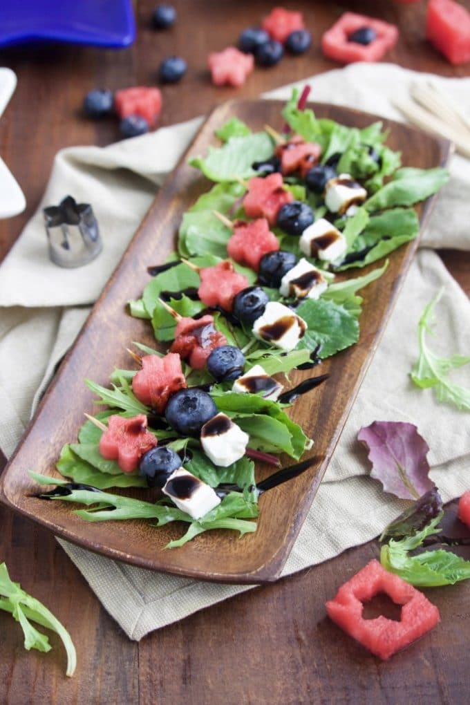 A wooden platter with red, white and blue skewers made with watermelon, blueberries and mozzarella cheese. A great 4th of July recipe.