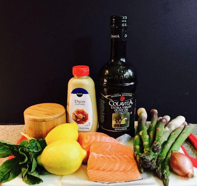 Fresh salmon fillets, mustard, lemon, olive oil, and a bunch of asparagus on a cutting board. 