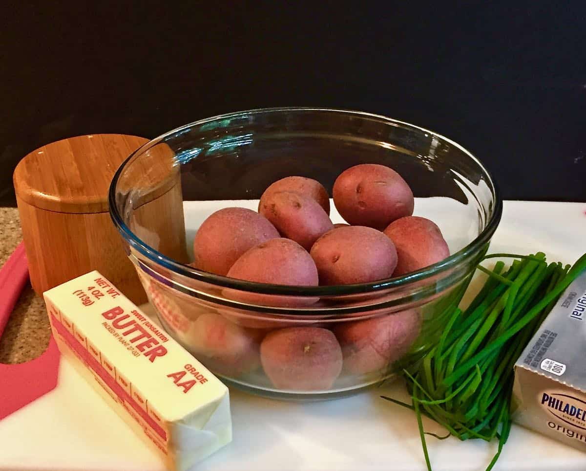 A bowl of red potatoes, a stick of butter, fresh chives and a box of cream cheese.