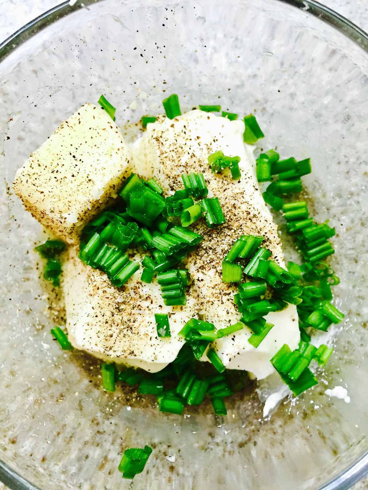 Chunks of cream cheese and butter in a clear glass bowl topped with chopped chives.