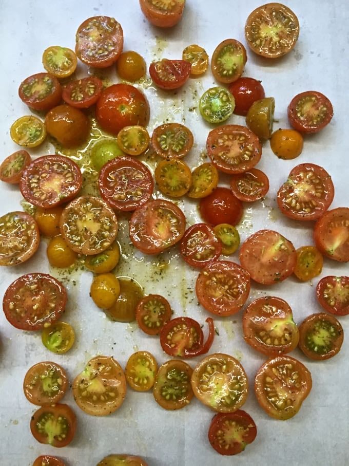 Sliced cherry tomatoes topped with olive oil and herbs on a baking sheet. 