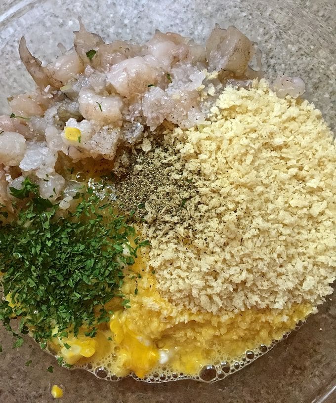 A clear glass bowl with shrimp, panko bread crumbs, eggs and parsley ready to mix. 