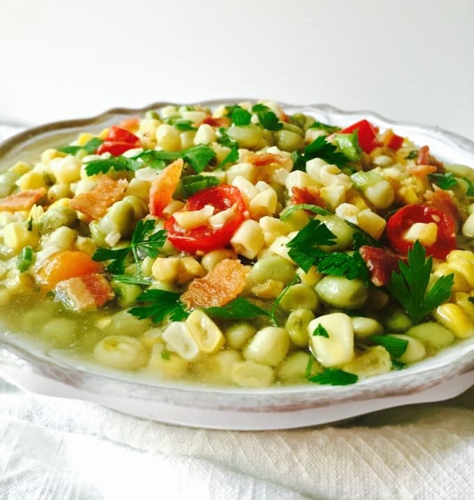  A bowl of corn, peas, and sliced tomatoes, topped with bacon and parsley. 