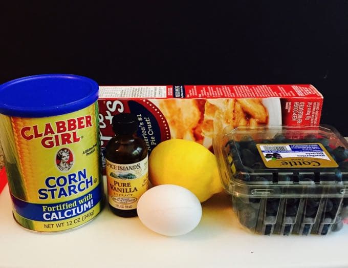 Ingredients corn starch, vanilla, blueberries, lemon, pie crusts and a lemon on a kitchen counter.