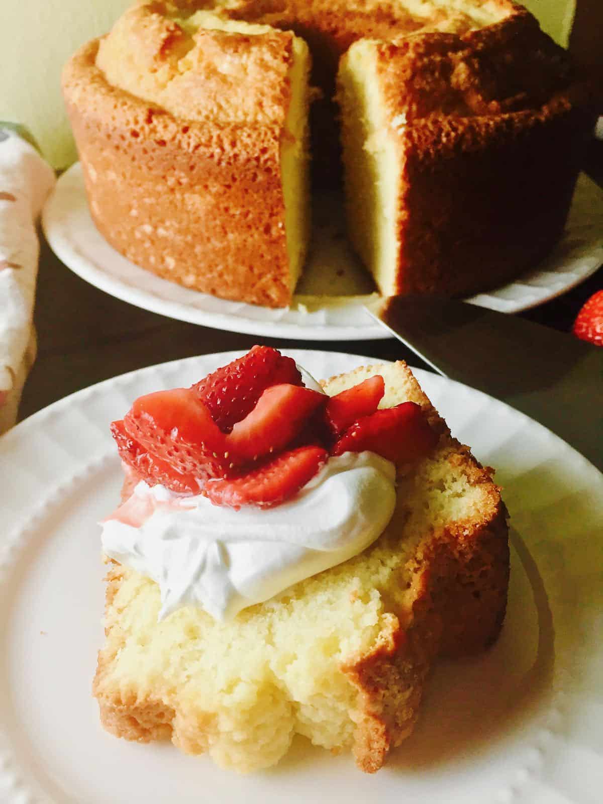 A large pound cake in the background with a slice cut out. The slice is topped with whipped cream and sliced strawberries. 