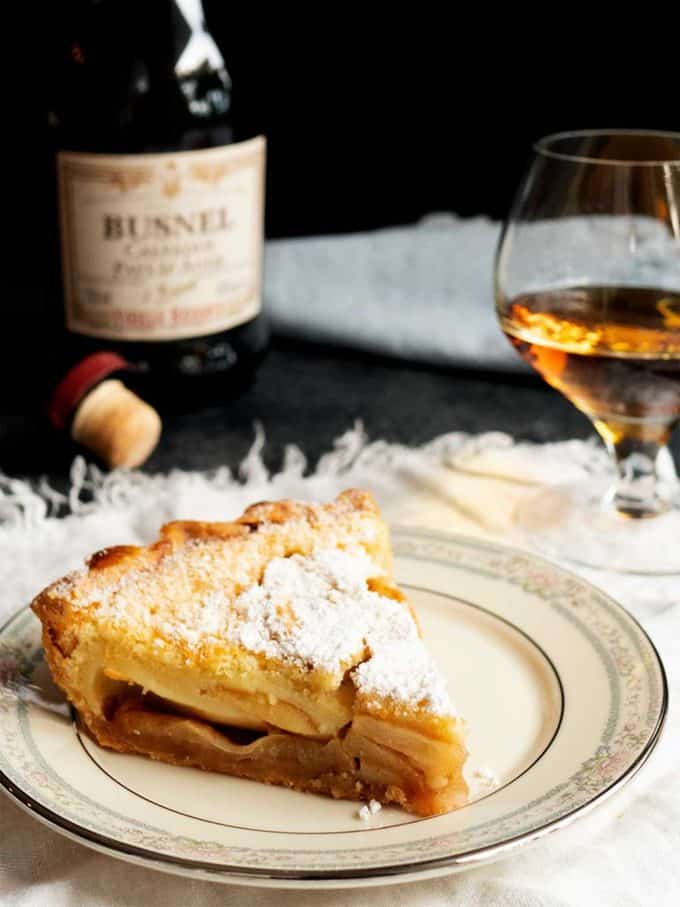 A slice of apple tart on a white plate with a glass of wine in the background.