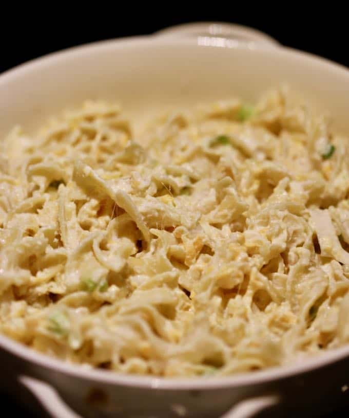 Mixing cooked egg noodles with cheese in a white casserole dish.