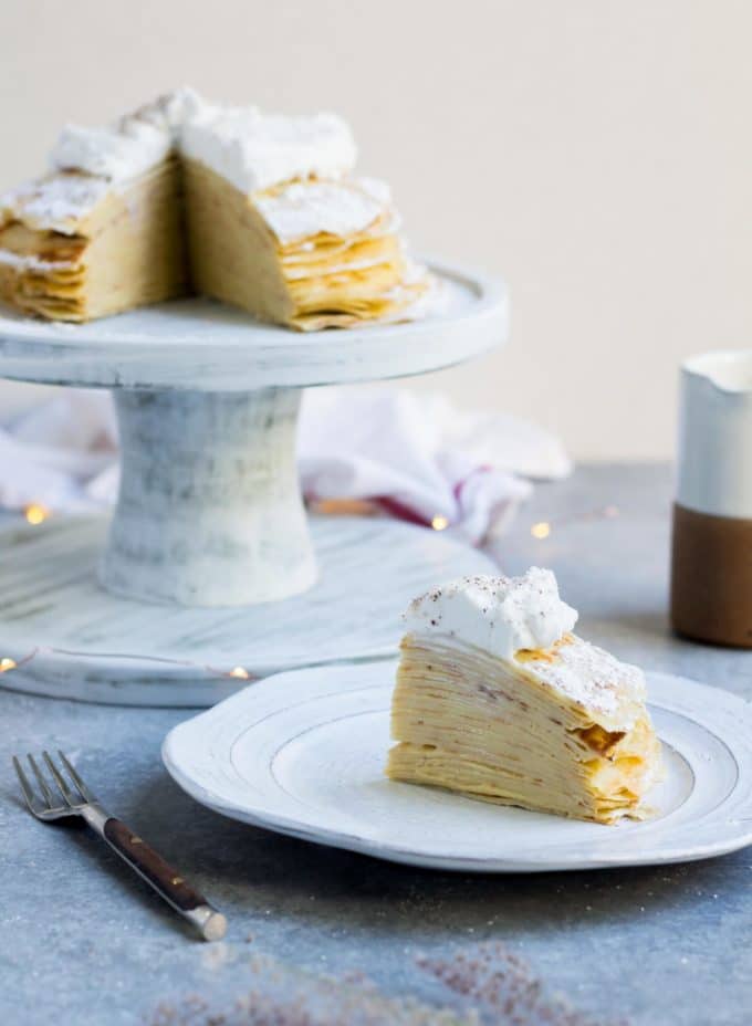 Eggnog Crepe Cake on a cake plate and a slice on a saucer. Both are topped with whipped cream. 