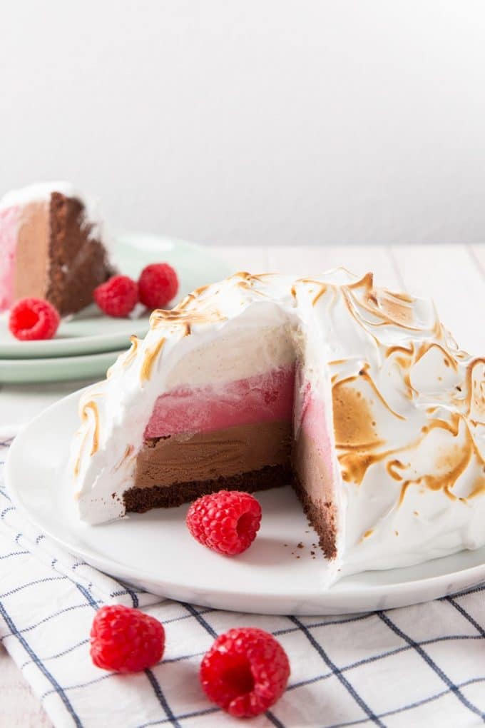 Baked Alaska on a plate with a slice cut out showing chocolate, strawberry and vanilla ice cream topped with a meringue. 