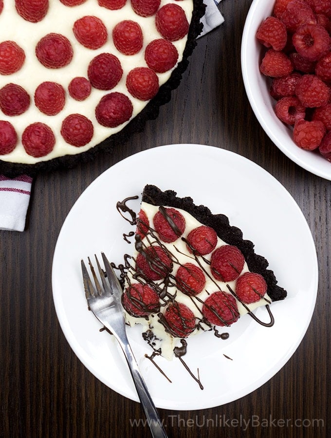 A slice of Raspberry White Chocolate Tart on a white plate topped with raspberries and a drizzle of chocolate.