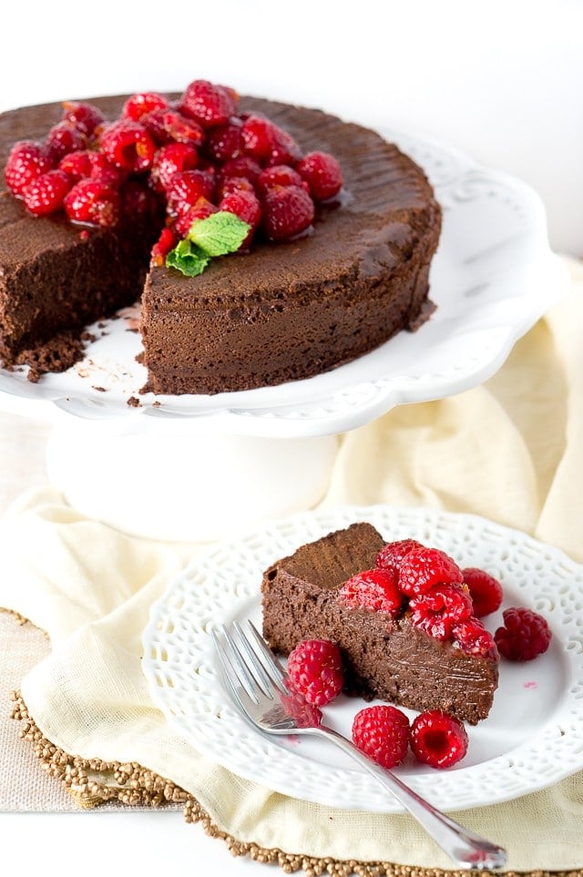 A Flourless Chocolate Cake on a cake platter, topped with raspberries and with a slice on a plate next to it. 