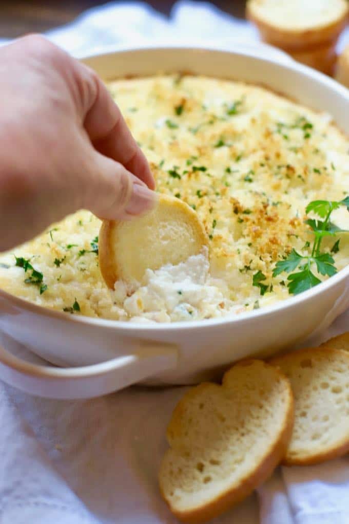 Dipping a toasted bread round into Hot and Cheesy Baked Shrimp Scampi Dip in a white baking dish