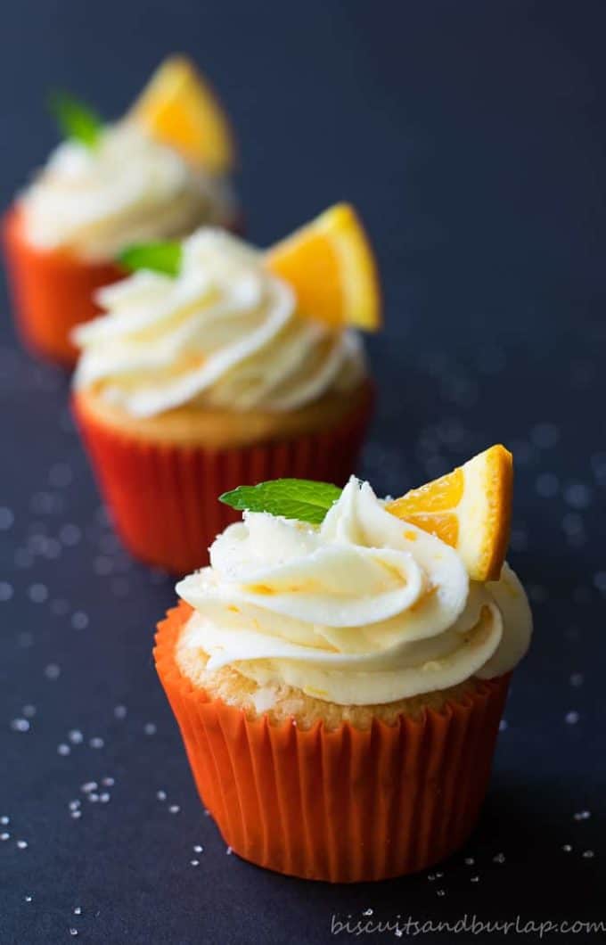 A row of orange cupcakes with orange cream cheese icing garnished with orange slices and mint leaves