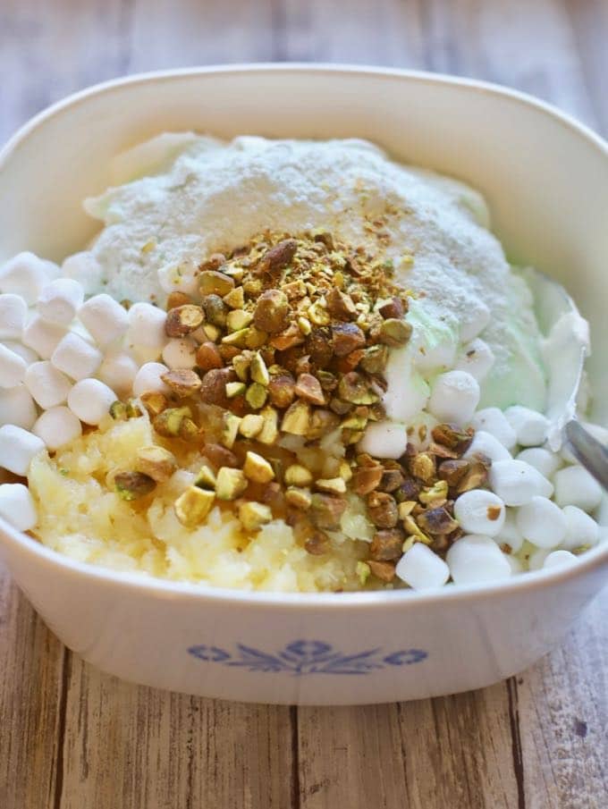 Marshmallows, pudding mix, pineapple and pistachios in a corning ware bowl.