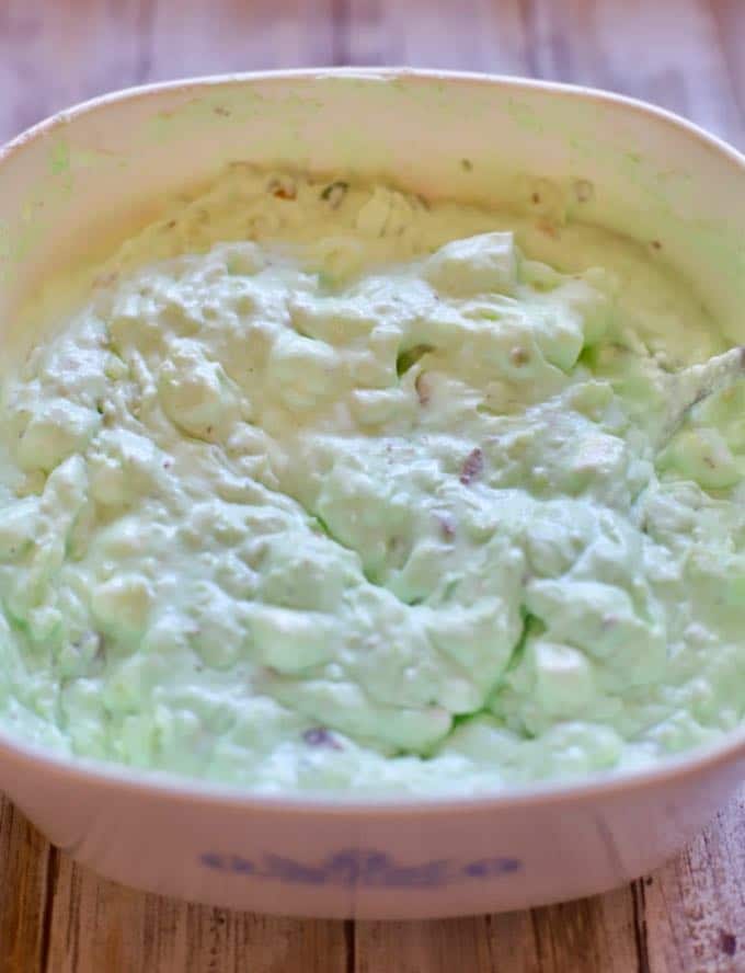 Cool Whip mixed with marshmallows, pineapple and pistachios in a white corning ware bowl.